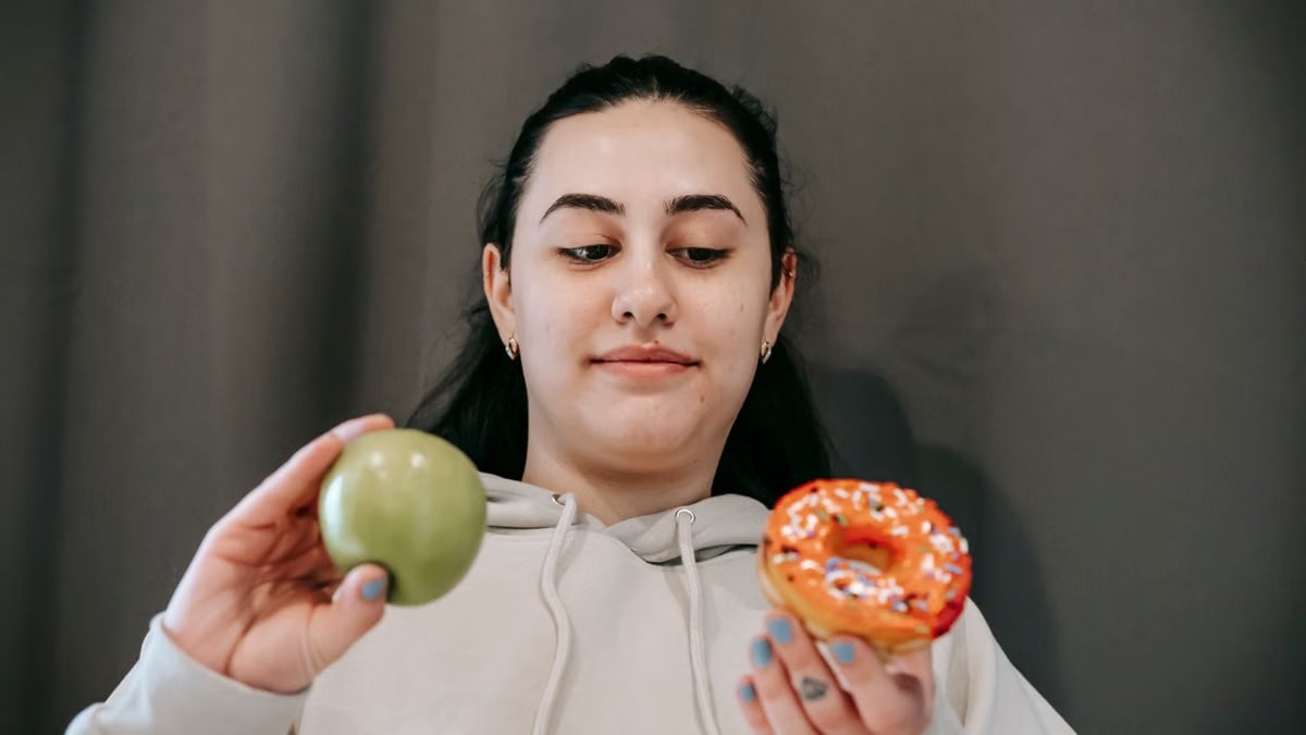 Content young female in hoodie deciding between sweet unhealthy doughnut and green ripe apple in light room